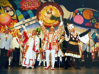 Kölner Dreigestirn der Session 1998 auf einer Bühne Das Kölner Dreigestirn der Session 1998 auf einer Bühne, im Hintergrund sieht man ein buntes Bühnenbild mit dem Gesellschaftslogo der Kölnischen Karnevals-Gesellschaft v. 1945