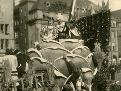Kölner Bauer und Kölner Jungfrau der Session 1930 Der Kölner Bauer und die Kölner Jungfrau auf einem Festwagen am Kölner Rosenmontag 1930, der Bauer reckt seinen Arm in den Himmel. Das Foto ist sepiafarben.