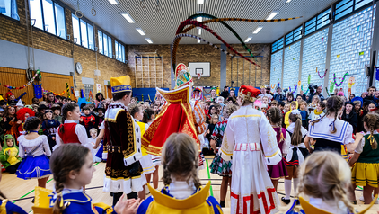 Das Kölner Kinderdreigestirn 2023 ist von hinten zu sehen, von links nach rechts Kinderbauer, Kinderprinz und Kinderjungfrau. Im Vordergrund sieht man die Pagen und Gardisten des Kölner Kinderdreigestirns in blau-gelben Uniformen. Im Hintergrund sind unzählige Kinder zu sehen, die tanzen und singen und jubeln. Das Foto wurde in einer Grundschulturnhalle aufgenommen.