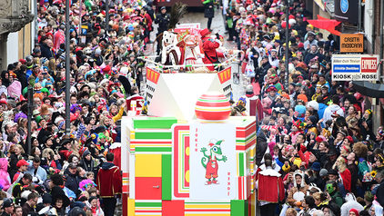 Man sieht von oben auf die Severinstraße direkt hinter der Severinstorburg herab. Rechts und links von der Straße stehen mehrere Reihen bunte Zuschauer, auf der Straße ist der Wagen des Kölner Kinderdreigestirns. Der Wagen ist bunt und mit dem Jecko geschmückt, man kann das Kölner Kinderdreigestirn sehen.