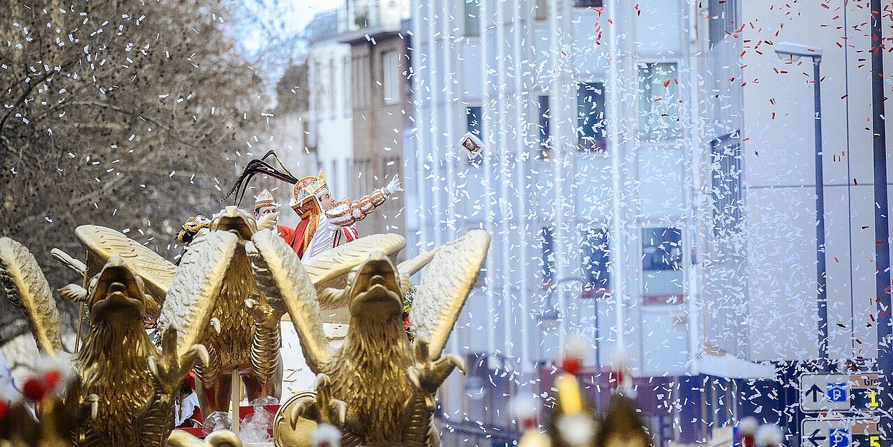 Prinz Karneval auf seinem Festwagen im Rosenmontagszug 2019, er wirft eine Schachtel Pralinen. Rund um den Wagen ist weißes Konfetti zu sehen und im Vordergrund sind die goldenen Fabelwesen zu sehen, die den Wagen des Prinzen ziehen.