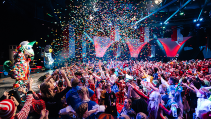 Zu sehen ist ein großer Raum mit einer Bühne und einem Zuschauerraum. Auf der Bühne tanz der Jecko, das Maskottchen vom Festkomitee Kölner Karneval. Vor der Bühne stehen unzählige Kinder in bunten Kostümen, die jubeln und tanzen und die Arme gehoben haben. In der Luft ist viel buntes Konfetti, dass auf die Kinder regnet. 